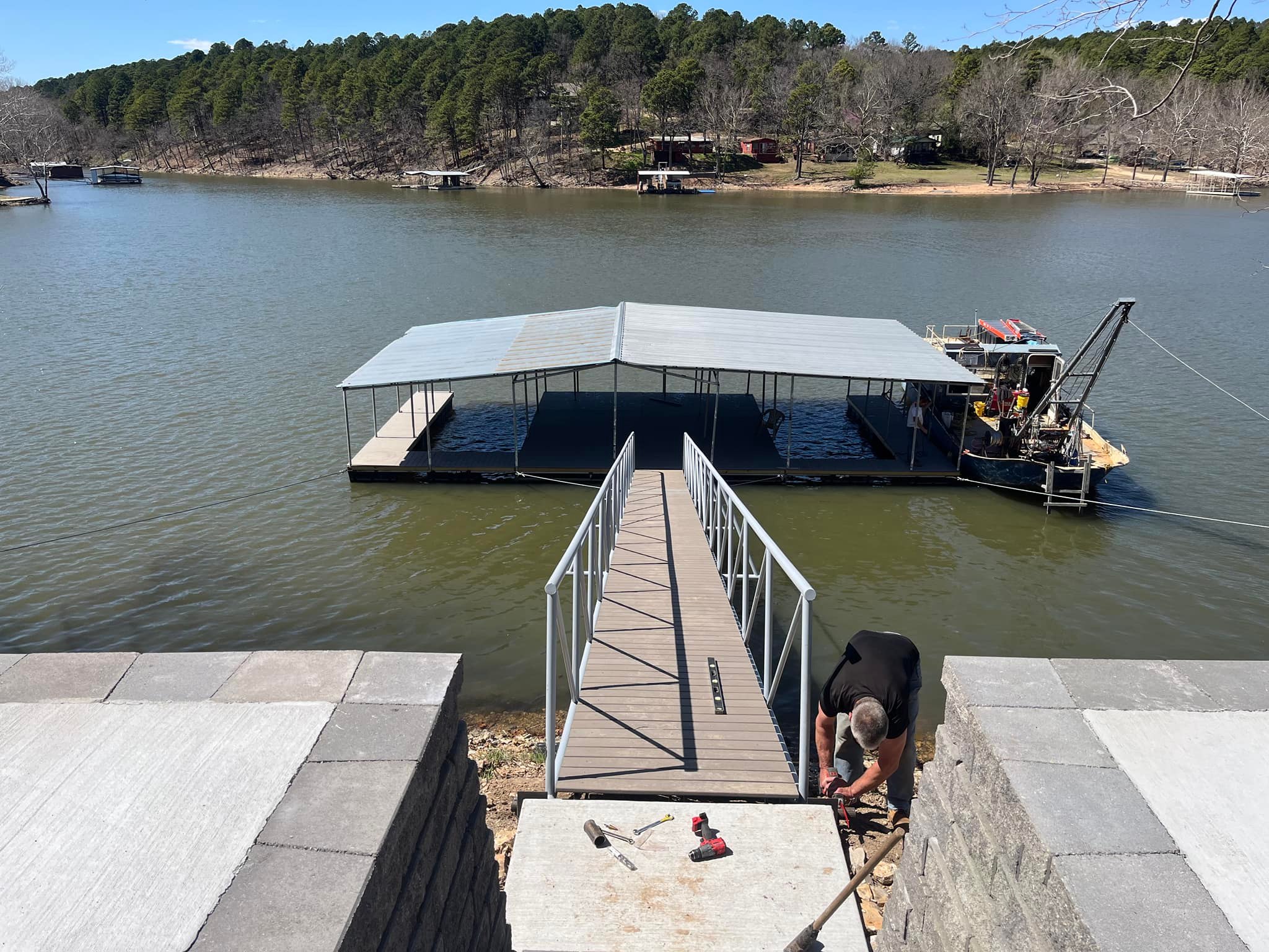 Covered floating dock installation at a lakeside property with a metal gangway leading from shore to the dock and construction equipment on the water