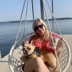 Woman sitting with a dog in a woven hanging chair on a Grand Lake dock overlooking calm water and the shoreline