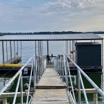 Covered boat dock with walkway and railings on Grand Lake in Oklahoma, featuring a metal roof, floating platform, and lakeside storage shed