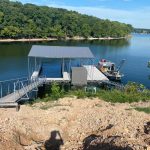 Covered multi slip dock with boat lift on Grand Lake Oklahoma, viewed from the shoreline with a gangway leading to the water and a workboat nearby