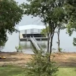 Covered lakeside dock with boat lift and walkway on Grand Lake in Oklahoma, viewed through trees from the shore